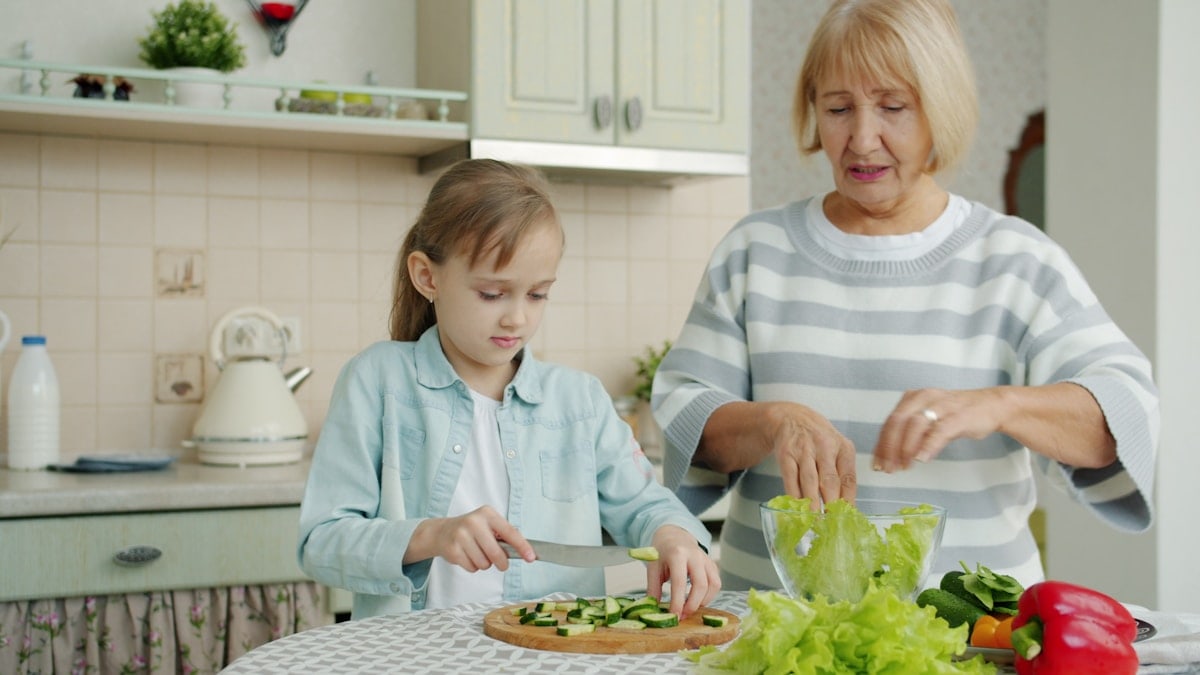 Grandma and cute child are cooking salad for lunch talking making healthy meal at home. Domestic activities, family life and happy childhood concept.